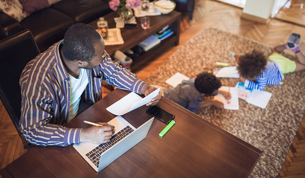 Photo of a father working from home in a living room while watching his children do schoolwork Photo of a father working from home in a living room while watching his children do schoolwork